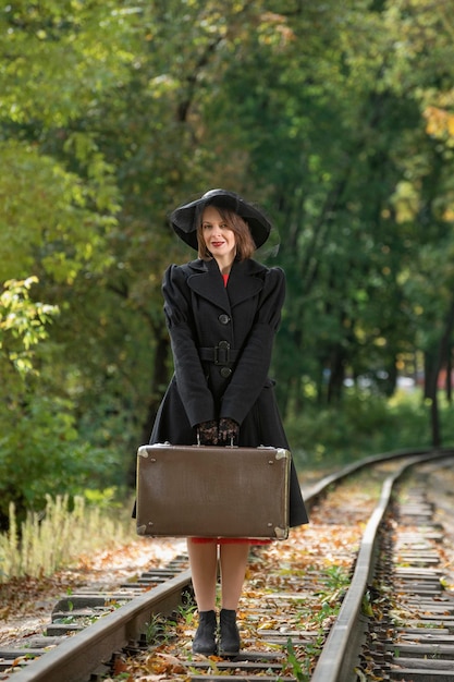 Vrouw in zwarte hoed en jas houdt retro koffer Madame vast in kleding van 3040s-stijl op mysterieuze wijze Verticaal frame