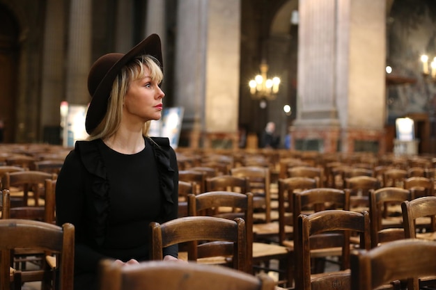 Vrouw in zwarte jurk en hoed zit alleen in de katholieke kerk. Bid tijd