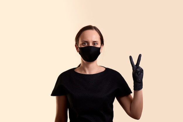 Foto vrouw in zwarte jurk gezichtsmasker en handschoenen overwinning hand over beige muur tonen