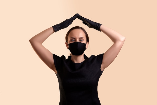 Vrouw in zwarte jurk gezichtsmasker en latex handschoenen met twee handen boven haar hoofd en een dak boven beige muur maken