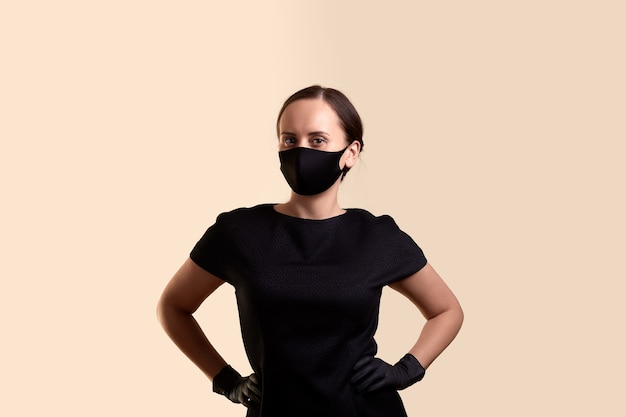 Vrouw in zwarte jurk gezichtsmasker en latex handschoenen pronken met zichzelf staande met armen over elkaar en over beige muur