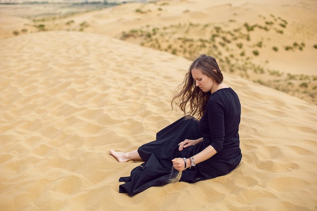Vrouw in zwarte lange jurk zit in de zomer met haar rug door de woestijnduinen
