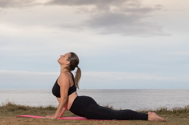 Vrouw in zwarte sportkleding doet pilates buiten op een roze mat met de zee