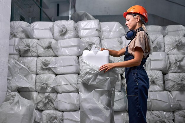 Vrouw industrier verpakking plastic vaten die in de fabriek bevatten, voorbereiding voor verkoop. jonge vrouw in blauw uniform en oranje veiligheidshelm. een heleboel verpakte jerrycans stapel flessen op de achtergrond