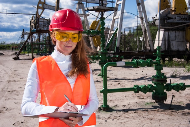 Vrouw ingenieur in gele bril op het olieveld met rode helm en werkkleding. Industriële site achtergrond.