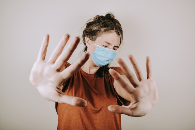 Vrouw is geschokt in een medisch masker en een bruin T-shirt toont een stopsignaal met haar handen geïsoleerd op een witte muur. De vrouw moedigt aan om thuis te blijven.