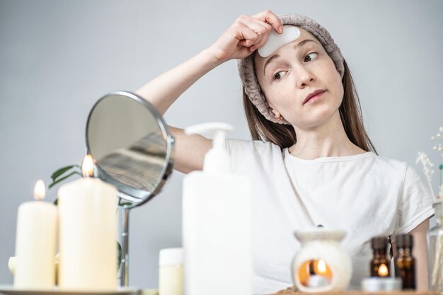 Vrouw kijkt in de spiegel en doet een massage met een gouachekrabber kaarsen en een aromalamp met essentiële oliën op tafel concept gezichtsverzorging aromatherapie ochtend- en avondrituelen