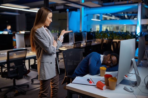 Vrouw kijkt op slapende manager, nachtkantoor levensstijl. vermoeide mannelijke personen op laptop, donker interieur, moderne werkplek