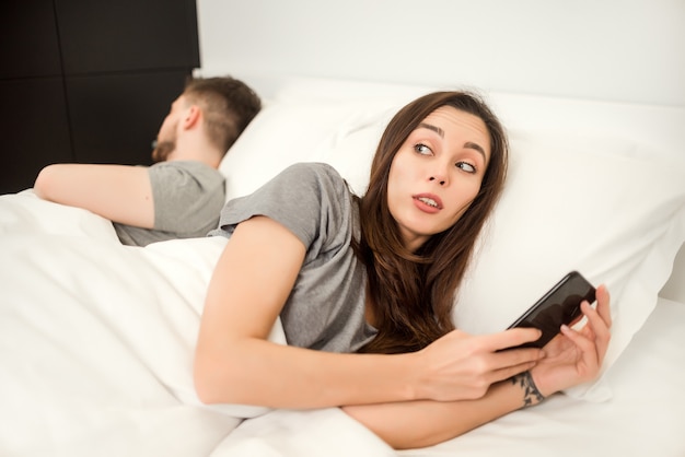 Vrouw kijkt stiekem naar haar telefoon terwijl man slaapt in bed