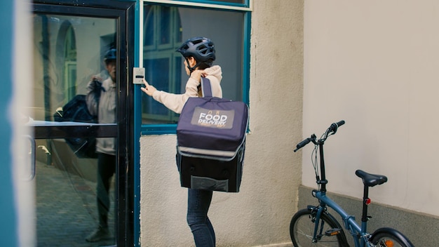 Vrouw koerier bezorgt fastfood pakket in papieren zak aan klant, restaurant afhaalmaaltijden voor de ingang van het kantoorgebouw. Horecamedewerker bezorgt maaltijdbestelling, medewerker op de fiets.