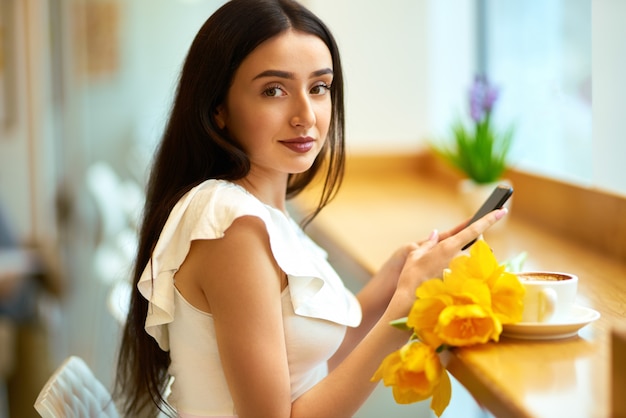 Vrouw koffie drinken en telefoon gebruiken. lente stijl. brunette vrouw In het mooie voorjaar. Mooie jonge vrouw met de lentetulpen bloeit boeket bij koffie. prachtige lentestemming