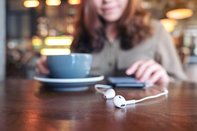 vrouw koffie drinken tijdens het gebruik van mobiele telefoon om muziek te luisteren met oortelefoon op houten tafel in café