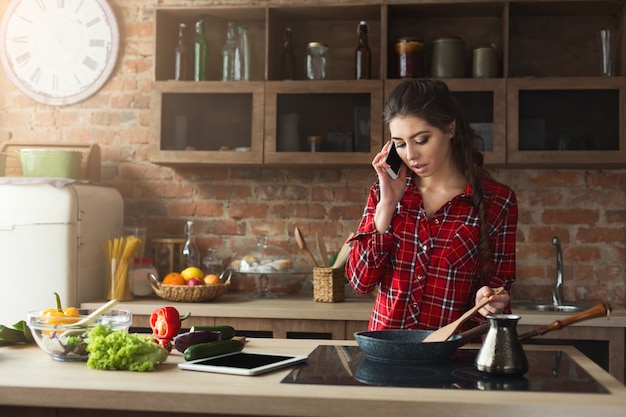 Vrouw koken gezond eten en praten op smartphone in de loft keuken thuis op zonnige dag. Groentesalade bereiden.