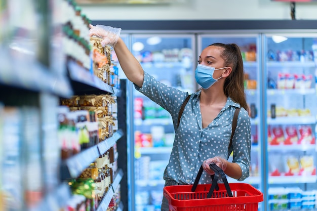 Vrouw koper in een beschermend gezichtsmasker en transparante handschoenen met winkelmandje kiest voedingsmiddelen in een supermarkt