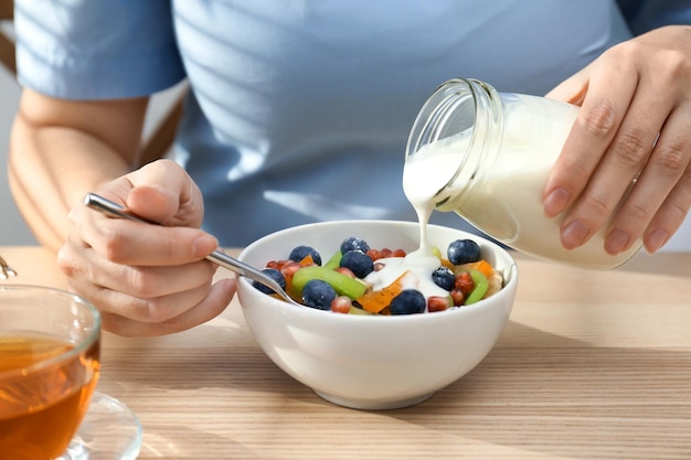 Vrouw lekkere yoghurt gieten op fruit in kom aan tafel