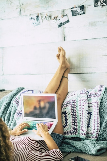 Vrouw liggend op de bank met benen die op de muur rusten, ontspant terwijl ze naar de computer kijkt. kijken naar streaming films of werken in een alternatieve kantoor- en lichaamshouding. vrouwen thuis met technologie