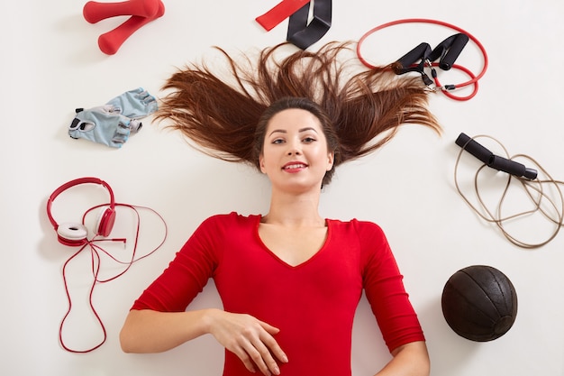 Vrouw liggend op de vloer na het trainen met halter, springtouw, fitness handschoenen, expander, elastische band, ziet er gelukkig en gezond uit, vrouw draagt rode kleding, sportief meisje op witte ondergrond.