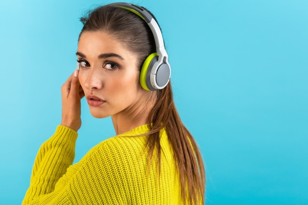 vrouw luisteren naar muziek in draadloze koptelefoon gelukkig dragen gele gebreide trui poseren op blauw