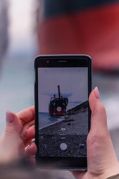 Vrouw maakt foto's van een groot schip op haar mobiele telefoon terwijl ze langs het strand loopt