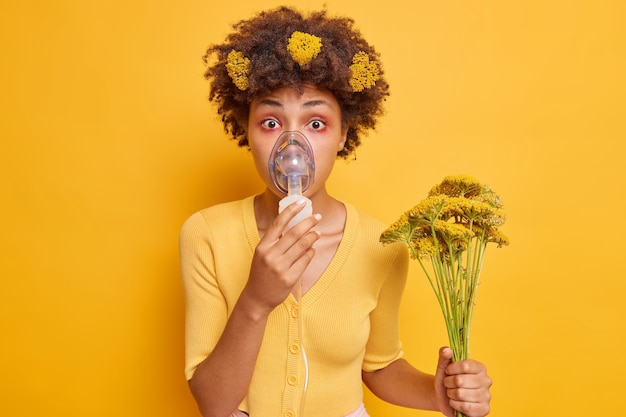 vrouw maakt inhalatievernevelaar draagt zuurstofmasker heeft ademhalingsmoeilijkheden door allergie houdt boeket met wilde bloemen vast draagt gele trui poseert binnen