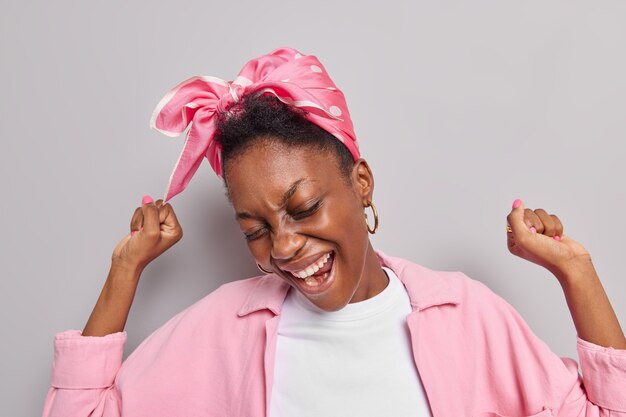 vrouw maakt kampioensdans steekt handen omhoog lacht gelukkig heeft plezier dansen op muziek draagt stijlvolle roze jas hoofddoek vastgebonden op hoofd geïsoleerd op grijs