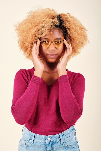 Vrouw met afro-kapsel die een zonnebril draagt en serieus naar de camera kijkt terwijl ze op een geïsoleerde achtergrond staat.