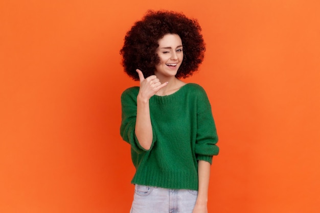 Vrouw met Afro-kapsel met vingers in de vorm van een telefoon in de buurt van het hoofd, telefonisch communicerend, camera kijkend met een speelse glimlach. Indoor studio opname geïsoleerd op een oranje achtergrond.