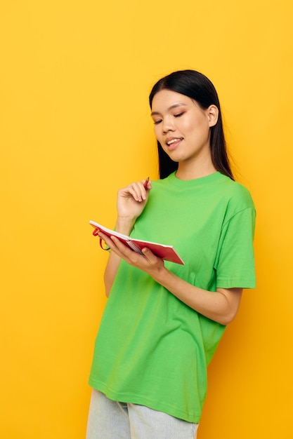 Vrouw met Aziatisch uiterlijk groene t-shirts met rood notitieboekje en pen gele achtergrond ongewijzigd
