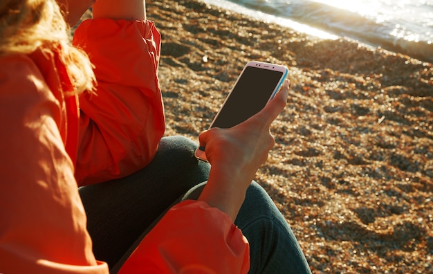 Vrouw met behulp van een smartphone zittend op het strand bij zonsondergang.