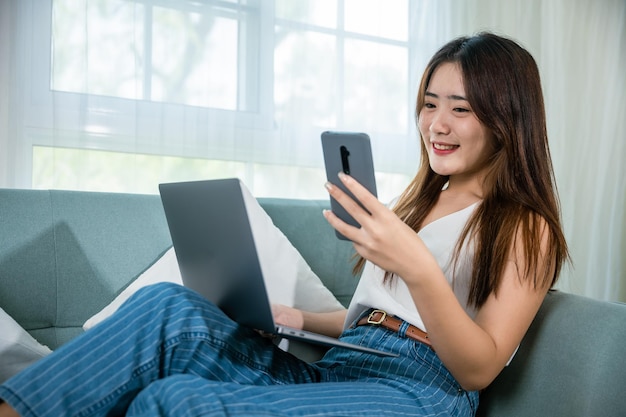 Vrouw met behulp van slimme mobiele telefoon zittend op de bank in de woonkamer thuis met laptopcomputer