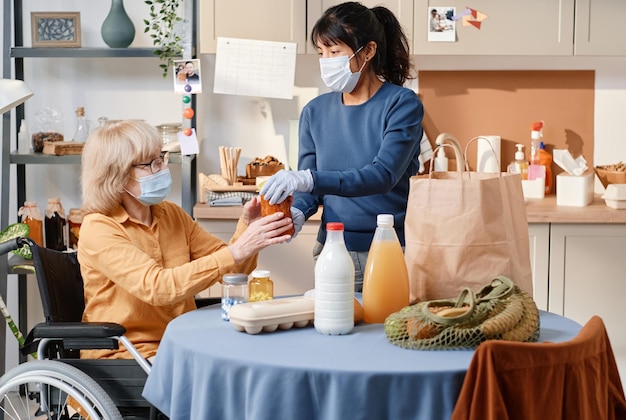 Vrouw met beschermend masker en handschoenen die voedsel aan huis levert voor oudere vrouw die rolstoel gebruikt