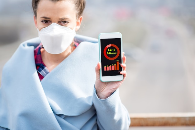 Vrouw met beschermend masker met smartphone met luchtvervuilingsmeting van PM10 buitenshuis