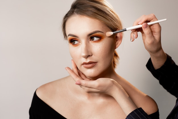 Vrouw met blote schouders die op lichte studio stellen. De handen van de visagist corrigeren make-up met een speciale borstel. Professionele make-up