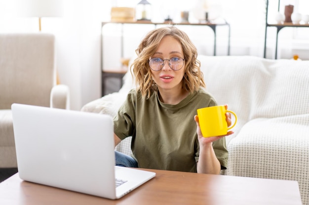 Vrouw met bril gebruik laptop drinken koffie in de ochtend zittend op de vloer in de buurt van de bank groot raam achtergrond interieur Freelance vrouw werken thuis Afstandsonderwijs student ontspannen kijken naar video lessen