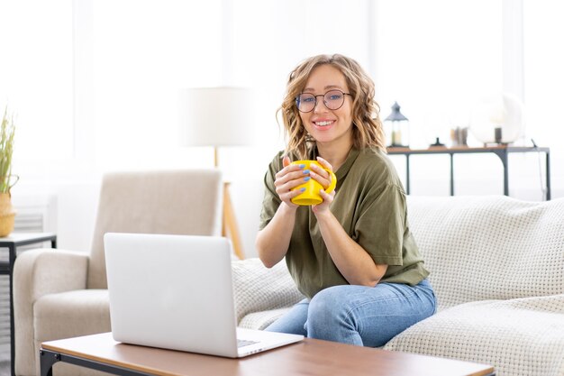 Vrouw met bril gebruik laptop typen toetsenbord zittend Bank groot raam achtergrond interieur Freelance vrouw werken vanuit huis Afstandsonderwijs student ontspannen kijken lessen videoconferentie