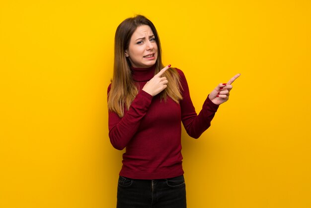 Vrouw met coltrui over gele muur bang en wijzend naar de kant