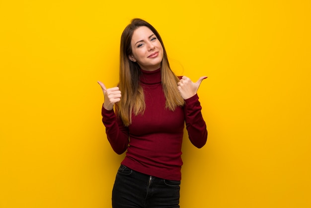 Vrouw met coltrui over gele muur die duimen op gebaar en het glimlachen geeft