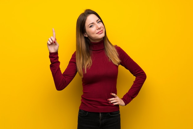 Vrouw met coltrui over gele muur die en een vinger in teken van het beste toont opheft