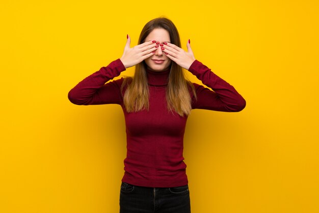 Vrouw met coltrui over gele muur die ogen behandelen door handen.