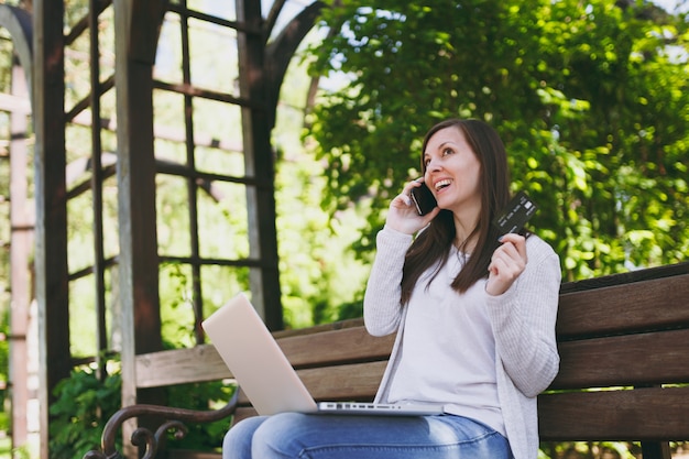 Vrouw met creditcard, praten op mobiele telefoon, met plezier een gesprek. vrouw zittend op een bankje bezig met moderne laptop in straat buiten. mobiel kantoor. freelance bedrijfsconcept.