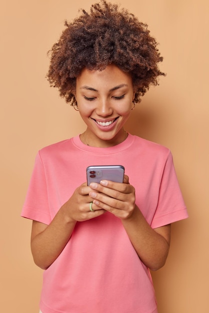 Foto vrouw met cury borstelig haar gebruikt mobiele telefoon chats online stuurt sms-berichten bladert sociale netwerken draagt casual t-shirt geïsoleerd op beige. technologie concept