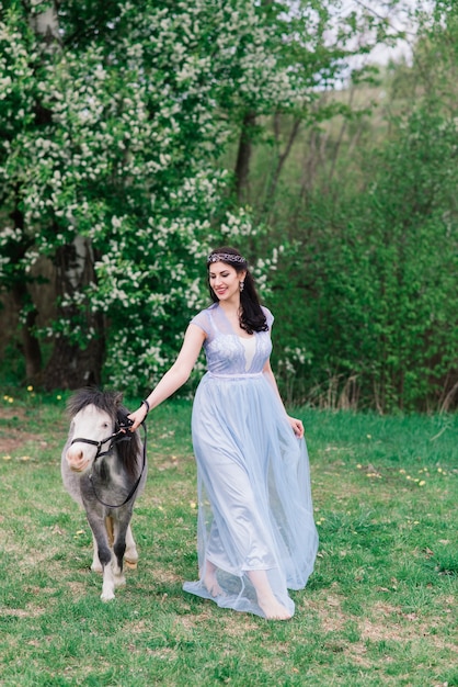 Vrouw met donker haar in een mooie jurk loopt met een grijze pony door het bos