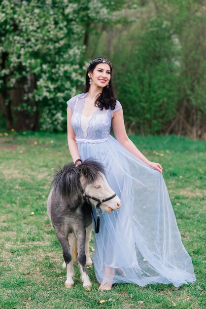 Vrouw met donker haar in een mooie jurk loopt met een grijze pony door het bos