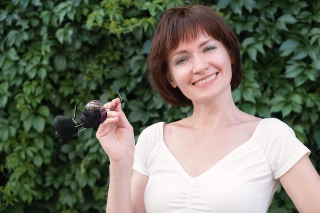 Vrouw met donkerrood haar staat en glimlacht voor een muur bedekt met bladeren