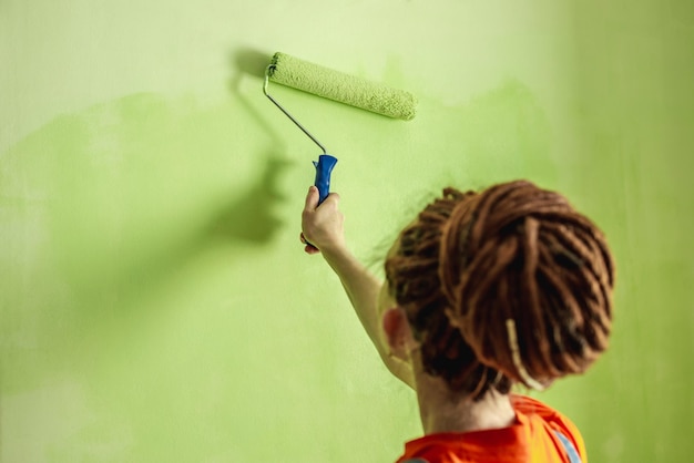 Vrouw met dreadlocks in oranje kleding schildert de muur met een roller in groene kleur Concept van reparatie renovatie van het nieuwe appartement Kopieer ruimte en close-up