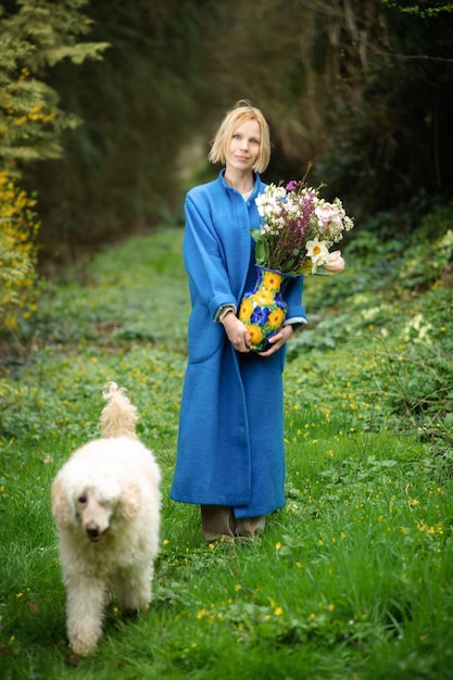 Vrouw met een boeket en een hond in het bos Blauwe jas en groene bosachtergrond Wandelgemak van het natuurbos