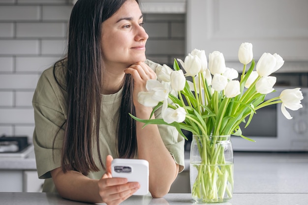 Vrouw met een boeket witte tulpen gebruikt thuis een smartphone in de keuken