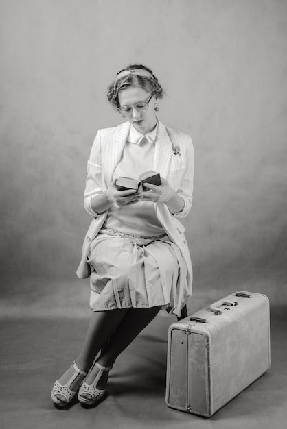 Foto vrouw met een bril die op een stoel zit met een koffer aan haar voeten die boek leest