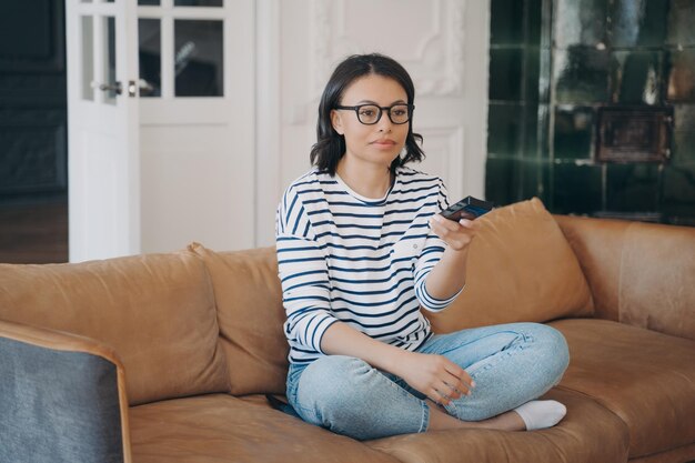 Vrouw met een bril die van televisiezender verandert en tv kijkt terwijl ze thuis op de bank zit