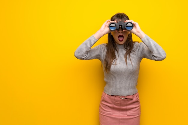 Vrouw met een bril over gele muur en kijkt in de verte met een verrekijker